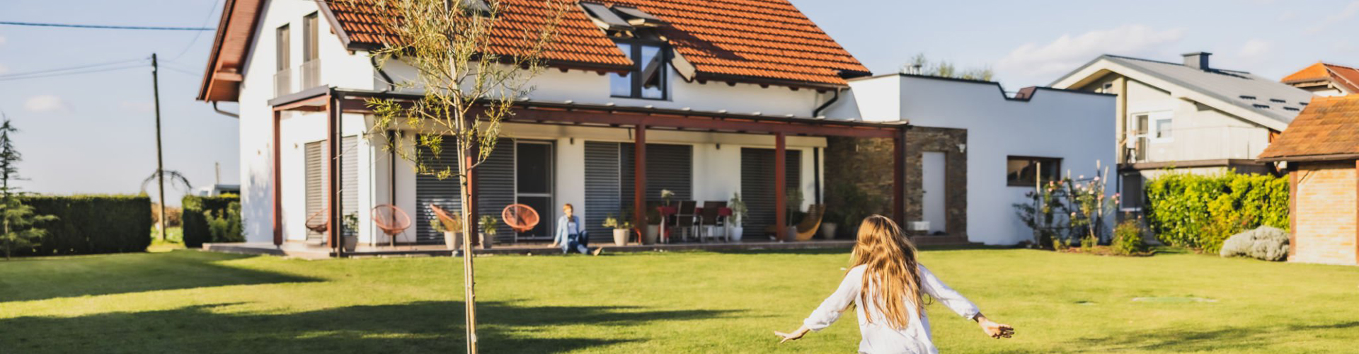 Little girl with long brown hair is running through the garden on lawn with a tree next to her,spreading her arms while running,house in the background,during summertime,rear view